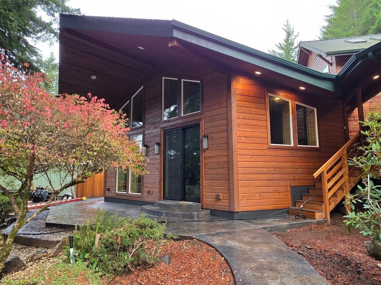 Modern wooden cabin with large windows, black stone path, and red shrubs in a wooded setting.