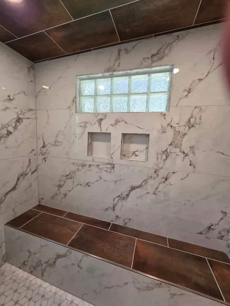 Empty tiled shower with marble-look walls, dark brown bench and ledge, and small frosted glass blocks.