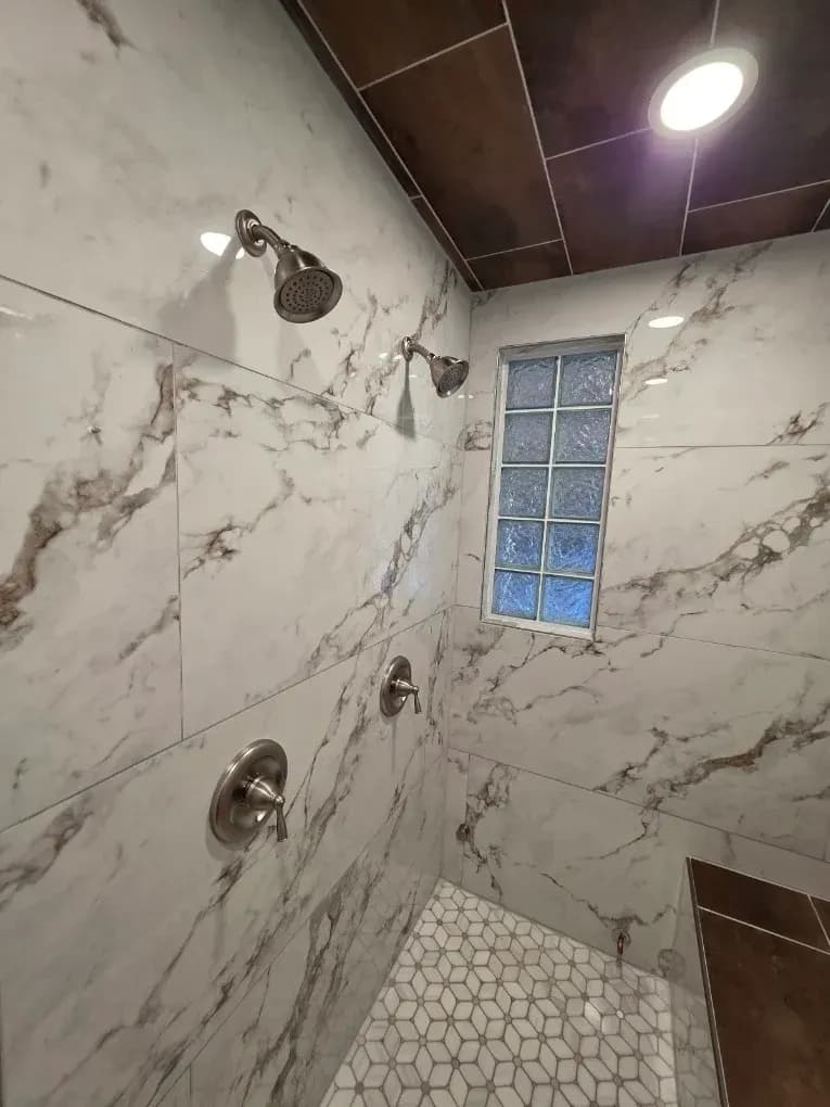 Empty shower stall with marble-look tile, chrome fixtures, and a small frosted window.