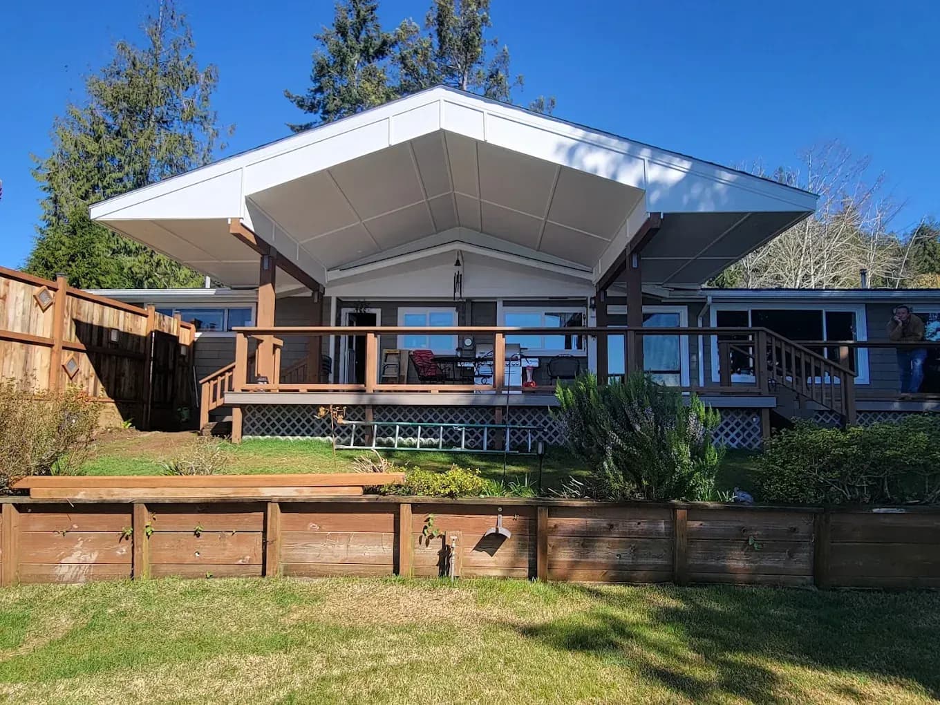 Modern house with a large white roof, wooden deck, and fenced backyard under a blue sky.