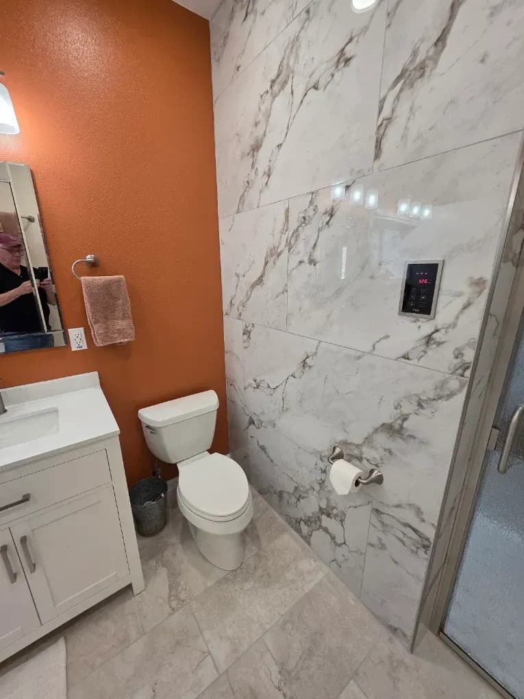 Small bathroom with white toilet, marble tile shower wall, orange accent wall, and vanity sink.
